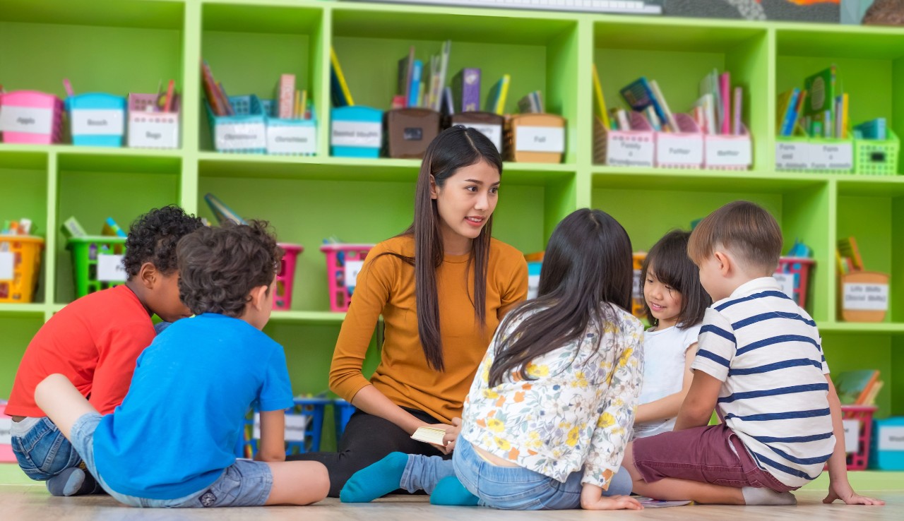 Asian female teacher teaching mixed race diversity group of kids reading book sitting on library floor in classroom,Kindergarten pre school concept